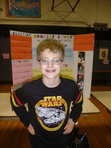 Zachary wearing a Star Wars shirt and standing in front of a school project with a smile