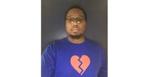 A 20-year old black man taking a selfie in front of a gray backdrop, with a light blue t-shirt on with a pink graphic heart 