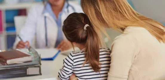 A mother and young daughter sitting at a desk across from a female doctor