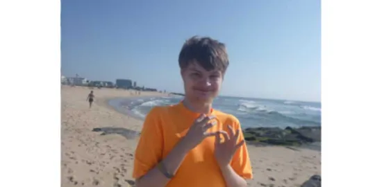 A young man in an orange shirt looks at the camera while standing on a sandy beach 