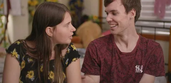 A white couple is smiling at each other mid laugh. The girl has a black shirt with sunflowers on it, and the boy has a red shirt. 