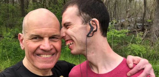 Stuart Spielman looking at camera smiling with arm around his son's shoulder. Zak smiles while leaning his face towards side of Stuart's head.