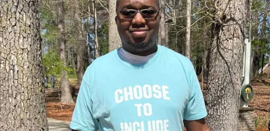 Young black man with glasses on stands in a teal t-shirt that says "Choose to Include" in white lettering 