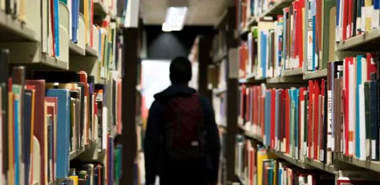 a student wearing a backpack and walking through an aisle of the school library