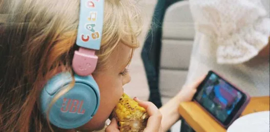 blonde toddler wearing headphones and looking at ipad while eating