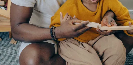 child wearing a yellow sweater and sitting on his dads lap playing with blocks