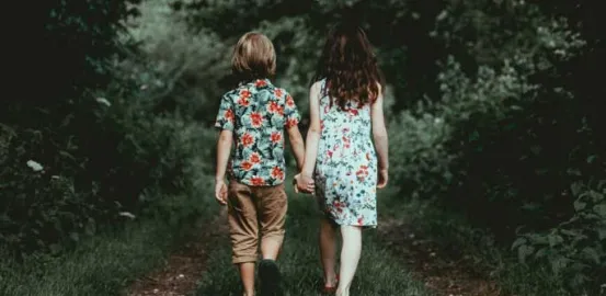 two children walking on a wooded path while holding hands