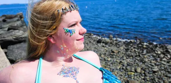 woman with blonde hair sitting by the ocean with blue gems on fer face