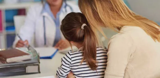 young girl sitting on her moms lap while speaking with a doctor at a desk