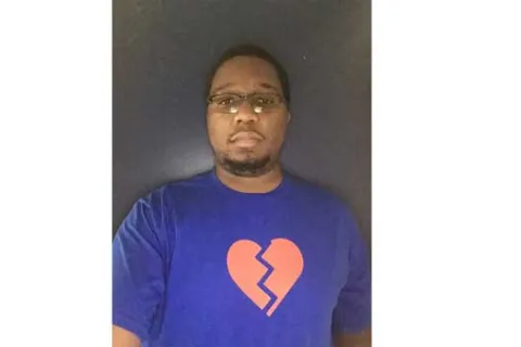 A 20-year old black man taking a selfie in front of a gray backdrop, with a light blue t-shirt on with a pink graphic heart 