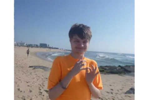 A young man in an orange shirt looks at the camera while standing on a sandy beach 