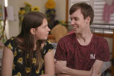 A white couple is smiling at each other mid laugh. The girl has a black shirt with sunflowers on it, and the boy has a red shirt. 