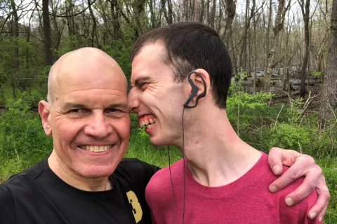 Stuart Spielman looking at camera smiling with arm around his son's shoulder. Zak smiles while leaning his face towards side of Stuart's head.