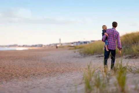 a man in a plaid shirt walking on the beach and carrying a child