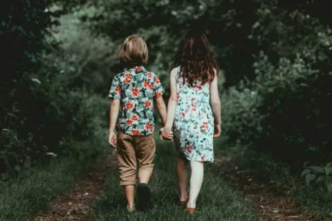 two children walking on a wooded path while holding hands