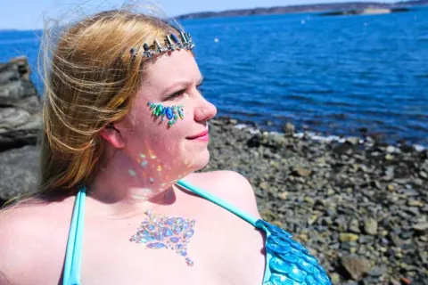 woman with blonde hair sitting by the ocean with blue gems on fer face