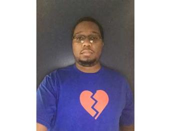 A 20-year old black man taking a selfie in front of a gray backdrop, with a light blue t-shirt on with a pink graphic heart 