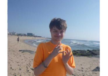 A young man in an orange shirt looks at the camera while standing on a sandy beach 