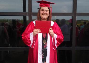 A girl with brown hair holding a diploma and wearing a red graduation cap and gown