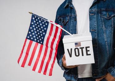 person holding a white box with "VOTE" on the front and an American flag hanging out the top