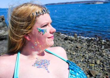 woman with blonde hair sitting by the ocean with blue gems on fer face