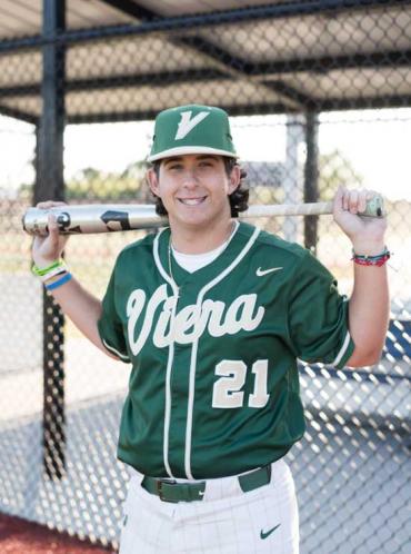 A teen in a green baseball jersey stands with a bat 