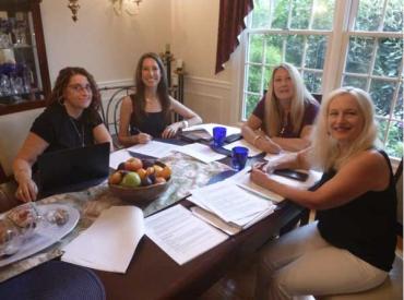 Four women sit on a wooden table and chair with stacks of papers between them, they are all smiling at the camera 