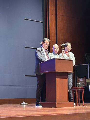 Keith Wargo, Stuart Spielman and Zak Spielman standing behind podium on stage. Zak is holding his phone up to his face