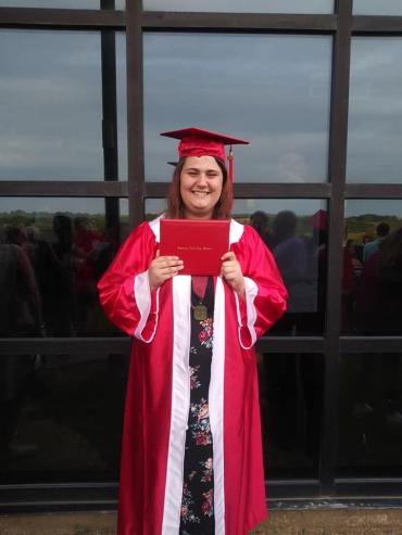 A girl with brown hair holding a diploma and wearing a red graduation cap and gown