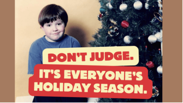 A young boy in a navy shirt sits next to a christmas tree with the words "don't jude. It's everyone's holiday season." 