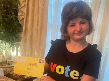 Self-advocate Kelsey N. wearing a tshirt that says vote and holding her mail in ballot envelope